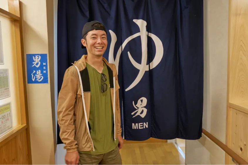 男湯ののれんの前で笑顔を見せるけんたさん
